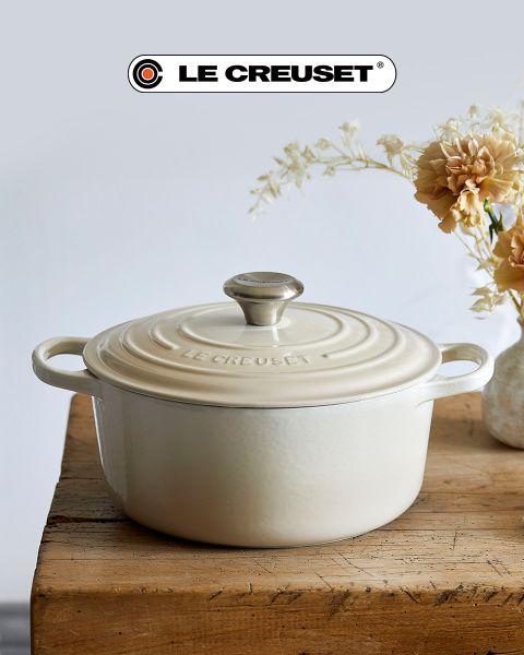 Eine Küchenszene mit einem roten Schmortopf, einer antihaftbeschichteten Bratpfanne, einem stapelbaren Topfset aus Edelstahl, einem schwarzen Kochtopf und einer Schüssel mit Kirschtomaten auf einer hellen Holztheke vor einer weißen Fliesenrückwand. Das Le Creuset-Logo ist sichtbar.