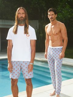 Ein Mann ohne Hemd in einem schwarzen Blazer und dunklen Shorts sitzt auf einem weißen Stuhl an felsigen Klippen am Meer und hält ein Glas Weißwein in der Hand, während im Hintergrund die Wellen rauschen.