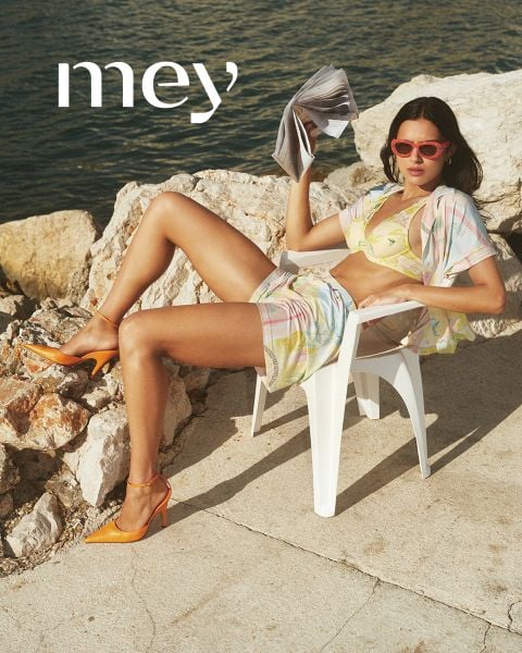 Ein Mann in schwarzen Boxershorts und eine Frau in einem weißen Tanktop und Unterwäsche stehen nebeneinander im Freien an einem Geländer, mit dem Meer und dem bewölkten Himmel im Hintergrund. Das Wort "mey" steht am unteren Rand.