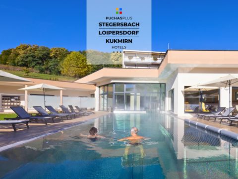 Zwei Menschen entspannen in einem langen Außenpool, der von Liegestühlen und Sonnenschirmen umgeben ist. Die Umgebung ist sonnig, mit einem modernen Hotelgebäude im Hintergrund und Bäumen auf einem Hügel. Ein Schild zeigt die Namen: Stegersbach, Loipersdorf, Kukmirn Hotels.