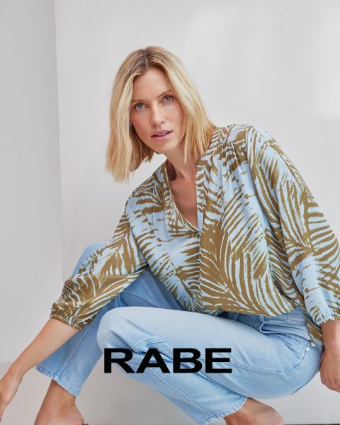 Eine Frau posiert vor einer weißen Wand und trägt eine rote ärmellose Weste über einer rot-weiß gestreiften Bluse und eine dunkle Hose. Am unteren Rand des Bildes ist das Wort "RABE" zu lesen.