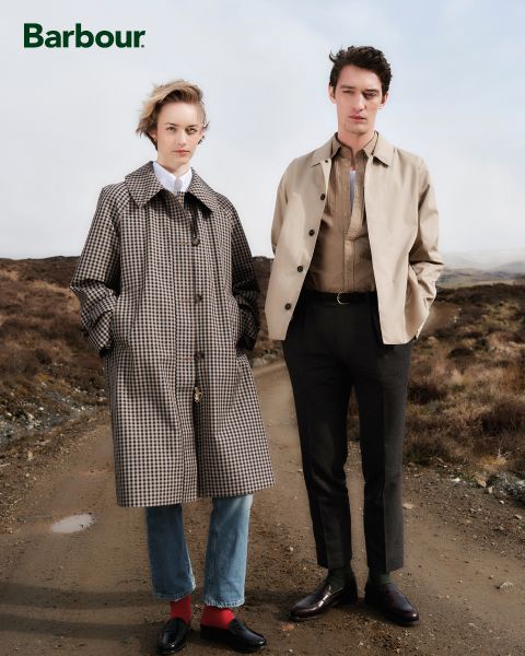 Eine Person in einem Mantel mit Hahnentrittmuster steht neben einer anderen Person, die eine beige Jacke, ein braunes Hemd und dunkle Hosen trägt. Sie befinden sich auf einem Feldweg in einer ländlichen Landschaft. Das Barbour-Logo befindet sich in der oberen linken Ecke.