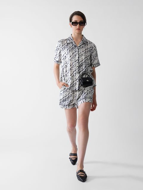 Eine Person mit Sonnenbrille, einem kurzärmligen, gemusterten Hemd und Shorts, schwarzen spitzen Schuhen und einer kleinen schwarzen Handtasche geht selbstbewusst vor einem schlichten weißen Hintergrund.