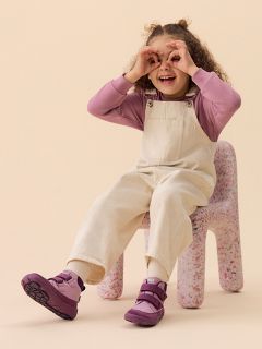 Ein lächelndes Kind mit grüner Mütze, gestreiftem Hemd und Weste sitzt auf einem pfirsichfarbenen Block vor einem gelben Hintergrund und trägt Stiefel und eine helle Hose.