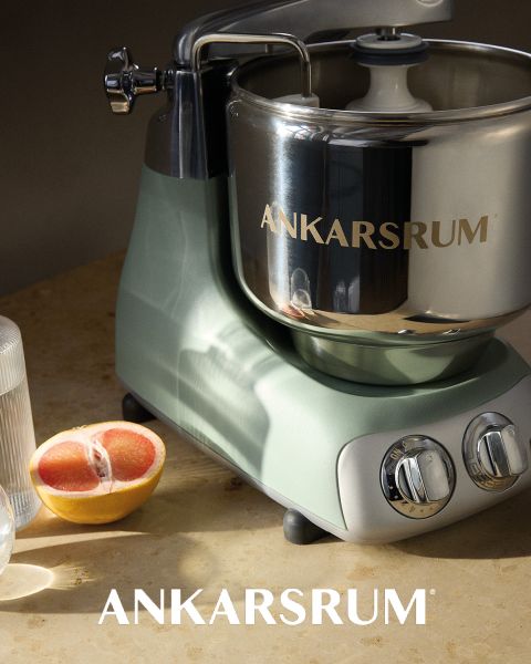 Eine glänzende Ankarsrum-Küchenmaschine mit einer Edelstahlschüssel und rosafarbenem Sockel wird auf einer dazu passenden rosafarbenen Oberfläche präsentiert, wobei der Markenname „ANKARSRUM“ sowohl auf der Schüssel als auch auf dem Hintergrund sichtbar ist.