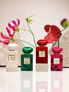 Acht Parfümflakons in verschiedenen Farben, darunter Rot, Grün, Blau, Rosa und Schwarz, sind auf beigen Podesten vor einem hellen Hintergrund angeordnet. Jeder Flakon trägt ein Etikett mit goldener Schrift.