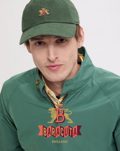 Eine Person mit kurzen, dunklen Haaren und Ohrring, die eine dunkle Jacke über einem weißen Hemd trägt, blickt in die Kamera. Die Innenseite der Jacke ist rot kariert. Auf der Vorderseite ist das Baracuta-Logo mit Löwe und „B“ abgebildet.