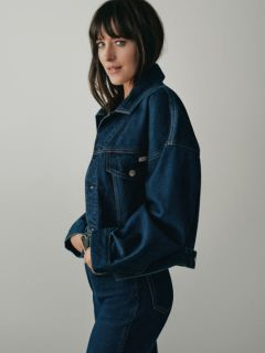 Eine Person mit langen blonden Haaren sitzt auf einer Holzbank und trägt eine blaue Jeansjacke, ein weißes Hemd und eine weit geschnittene Jeans. Im Hintergrund sind städtische Gebäude und Wolkenkratzer zu sehen.