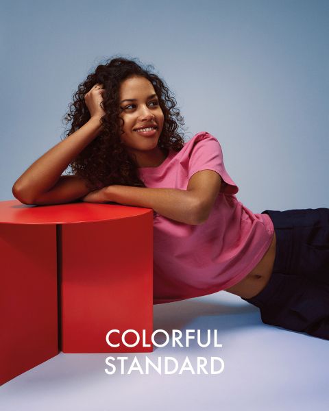 Eine Person mit langen blonden Haaren steht seitlich vor einem blauen Hintergrund. Sie trägt ein dunkelblaues T-Shirt, hellblaue Jeans und Turnschuhe. Der Text „COLORFUL STANDARD“ ist in Weiß dargestellt.