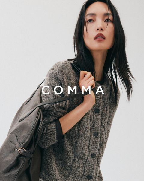 Eine Frau in einem karierten Mantel und goldenen Accessoires posiert mit einem hellbraunen und weißen Hund in einem passenden karierten Outfit. Das Wort "COMMA" erscheint in großer weißer Schrift quer über das Bild.