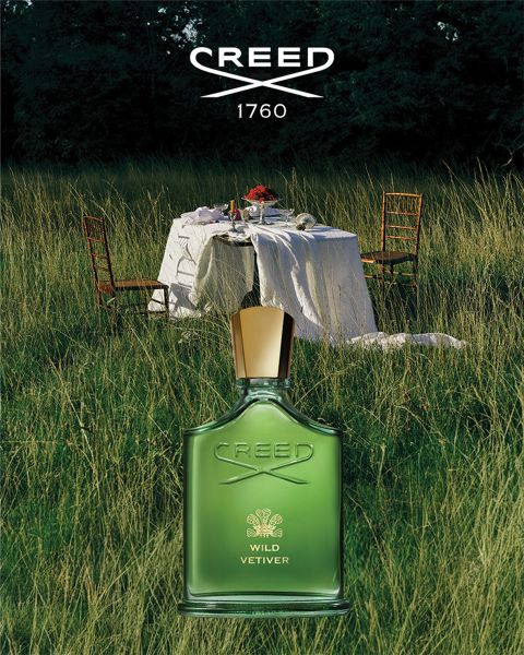 Ein weißer Creed Silver Mountain Water Parfümflakon liegt im Schnee mit verschneiten Berggipfeln im Hintergrund unter einem blauen Himmel.