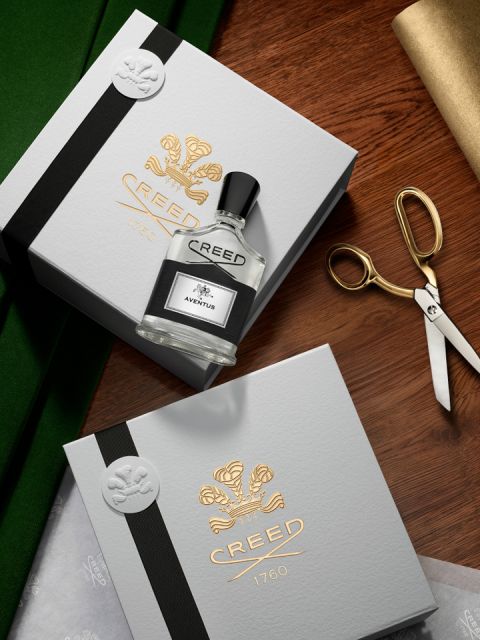 Eine Glasflasche mit Creed Aventus-Parfüm, umgeben von seinen Inhaltsstoffen: rote Beeren, weiße Blumen, Ananas, Moos und einige dunkle Gewürze. Der Hintergrund ist in einem sanften Beige gehalten.