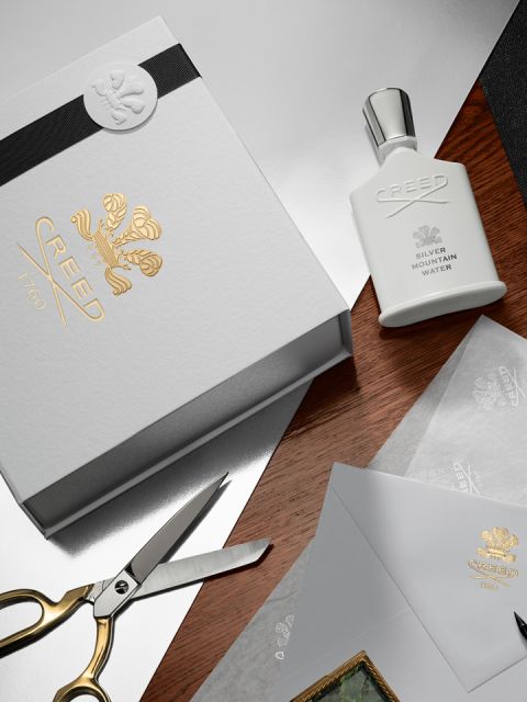 Eine luxuriöse Präsentation von Creed-Hautpflege- und Duftprodukten, darunter Flaschen, eine Tube und ein Stück Seife. Alle Artikel sind elegant auf einer neutral getönten Oberfläche mit weichem, fließendem Stoff im Hintergrund angeordnet.