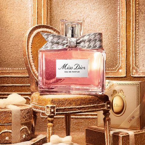 Eine durchsichtige Glasflasche mit „Miss Dior Eau de Parfum“, gefüllt mit rosa Flüssigkeit und einer silbernem, gemusterten Schleife um den Flaschenhals, vor einem schlichten weißen Hintergrund.