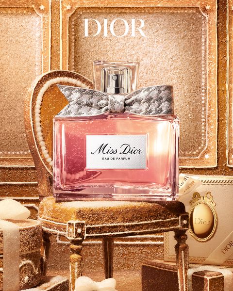 Ein Mann mit Tattoos und Schmuck lehnt sich an einem Tisch neben einer Flasche „Dior Sauvage Extrait“-Parfüm vor einer rauen Außenkulisse.