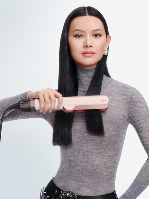 Frau mit langen blonden Haaren, die ein Glätteisen benutzt, ein weißes Oberteil trägt und nach vorne blickt. Kontrastreiches Schwarzweißbild.