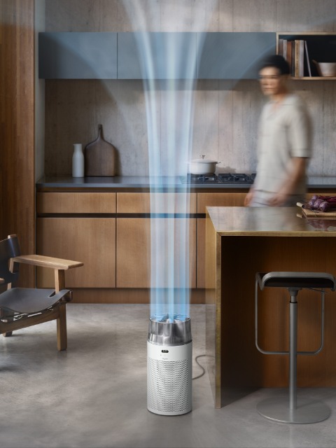 Eine Frau sitzt auf einem Stuhl und liest einem Kind in einem Kinderzimmer mit Kinderbett, Bücherregal und Lampe vor. Im Vordergrund stößt ein Luftreiniger in schlankem, modernem Design Luftströme aus. Der Raum hat helle Holzböden und sanfte blaue Wände.
