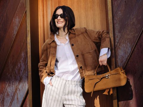 Eine Frau mit Sonnenbrille und brauner Wildlederjacke lächelt, während sie eine hellbraune Handtasche in der Hand hält. Sie trägt ein weißes Hemd und eine gestreifte Hose und steht vor einer Holztür.