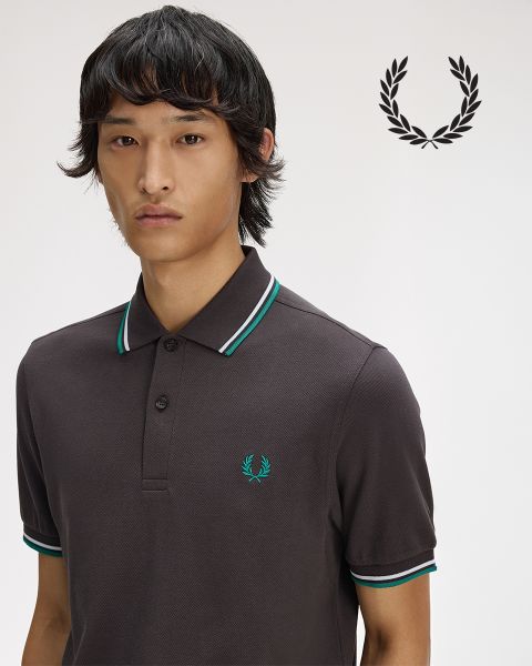 Eine Person mit langen geflochtenen Haaren blickt nachdenklich nach oben, während sie ein weißes Hemd mit Kragen zurechtrückt. Das Hemd ist mit einem Lorbeerkranz verziert. Das Bild ist schwarz-weiß.