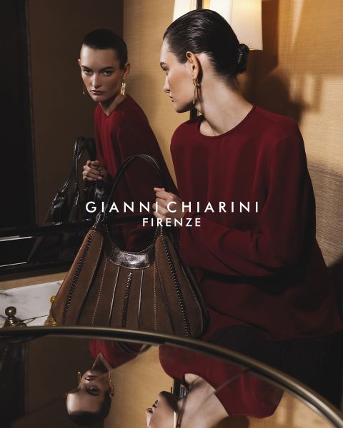Person, die einen langen schwarzen Trenchcoat aus Leder und eine Sonnenbrille trägt und eine kleine Tasche mit Leopardenmuster hält. Der Hintergrund ist grau, mit dem Text „GIANNI CHIARINI FIRENZE“ in Weiß über dem Bild.