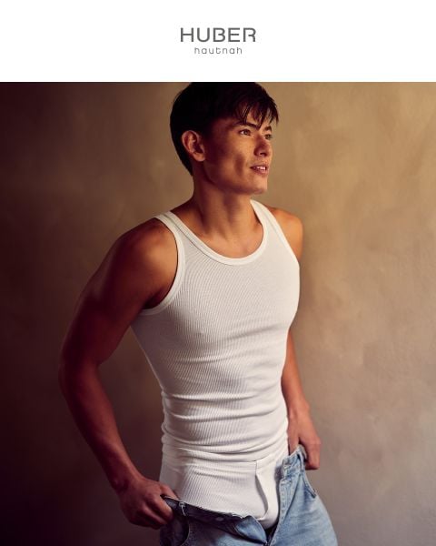 Ein lächelnder Mann in einem weißen gerippten Tanktop und Unterwäsche steht in einem Badezimmer mit warmer Beleuchtung, einem Spiegel und runden Lichtern hinter ihm. Der Text "HUBER hautnah" wird am unteren Rand des Bildes angezeigt.
