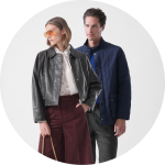 Zwei Personen stehen nebeneinander; die eine trägt eine braune Jacke, die andere eine Sonnenbrille und eine hellbraune Jacke über einem rosa Kleid mit kreisförmigen Mustern.