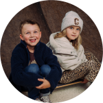Zwei lächelnde Kinder, ein Junge in einem gelben Hemd und ein Mädchen in einem gestreiften Hemd, sitzen dicht beieinander auf einer gelben Couch vor einem hellen Hintergrund.