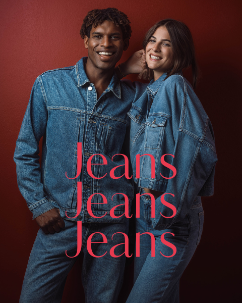Zwei Personen in blauen Jeansjacken und Jeans stehen lächelnd vor einer roten Wand. Über dem Bild ist in rosa Schrift der Schriftzug „Jeans Jeans Jeans“ zu sehen.
