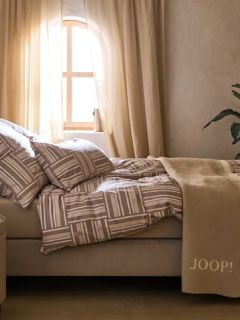 Ein minimalistischer Raum verfügt über eine beige Steinbank mit zwei strukturierten braunen und beigen Kissen, einer gemusterten Überwurfdecke und getrockneten Gräsern. Ein geflochtener Korb, eine rustikale Keramikvase und ein hoher Krug schmücken den Boden unter sanftem, natürlichem Licht.