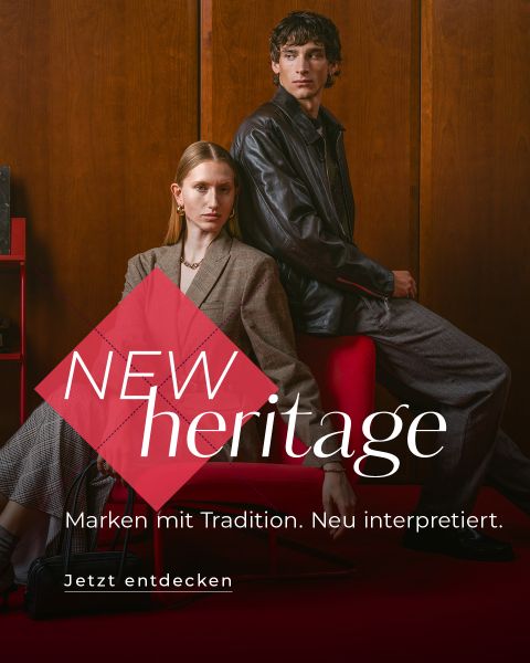 Eine Frau in einem Blazer sitzt auf einem roten Stuhl; hinter ihr steht ein Mann in einer Lederjacke. Text: „NEW heritage. Marken mit Tradition. Zeitlose Looks.“.