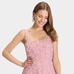 Lächelnde Frau in einem ärmellosen, hellrosa Kleid mit strukturiertem Rosenmuster, die Hand auf der Hüfte, mit funkelnden Tropfenohrringen vor grauem Hintergrund.