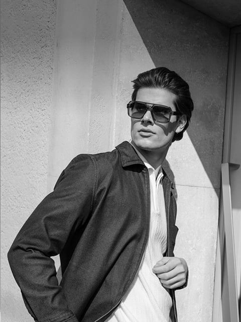 Ein Mann mit Sonnenbrille und Jacke steht draußen und schaut zur Seite, während das Sonnenlicht Schatten an die Wand hinter ihm wirft. Schwarz-Weiß-Foto.