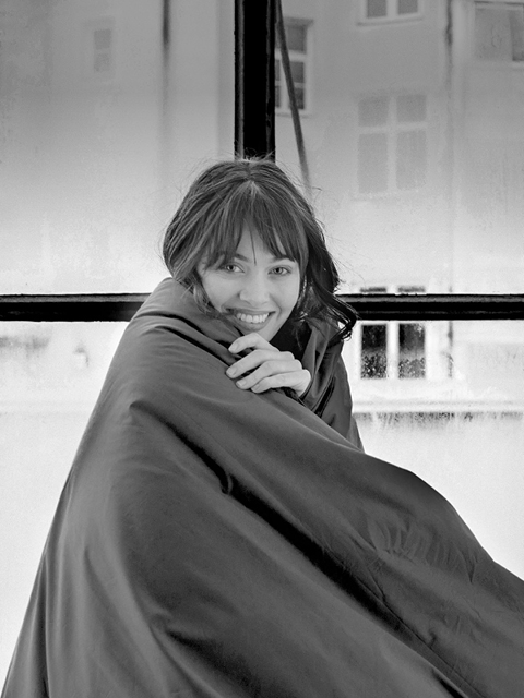 Eine lächelnde Frau, in eine Decke gehüllt, sitzt an einem großen Fenster, hinter dem an einem bewölkten Tag Gebäude zu sehen sind.