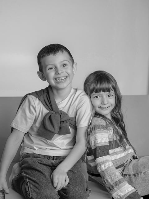 Zwei lächelnde Kinder, ein Junge und ein Mädchen, sitzen eng beieinander. Der Junge hat einen Pullover über die Schultern gelegt; das Mädchen hat lange Haare und trägt einen gestreiften Pullover.