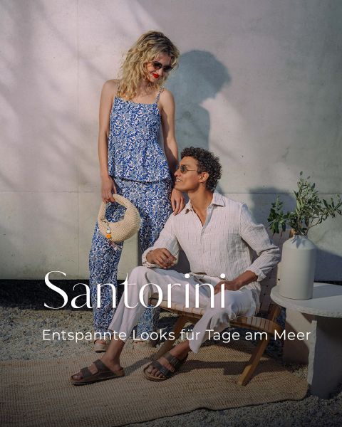 Eine Frau in einem blauen Blumenkleid steht neben einem sitzenden Mann in weißer Kleidung. Der Text lautet: "Santorini - Entspannte Looks für Tage am Meer.".