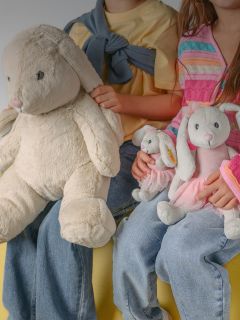 Ein lächelnder Junge mit blondem Haar sitzt in einem Haus und umarmt einen großen beigen Teddybär. Er trägt ein schwarz-weißes langärmeliges Hemd und eine helle Hose.