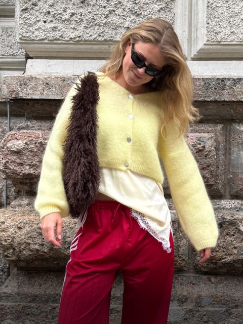 Eine Frau mit schwarzer Sonnenbrille, gelber Strickjacke, roter Hose und einer braunen, pelzbesetzten Umhängetasche steht vor einer strukturierten Steinmauer und lächelt leicht.