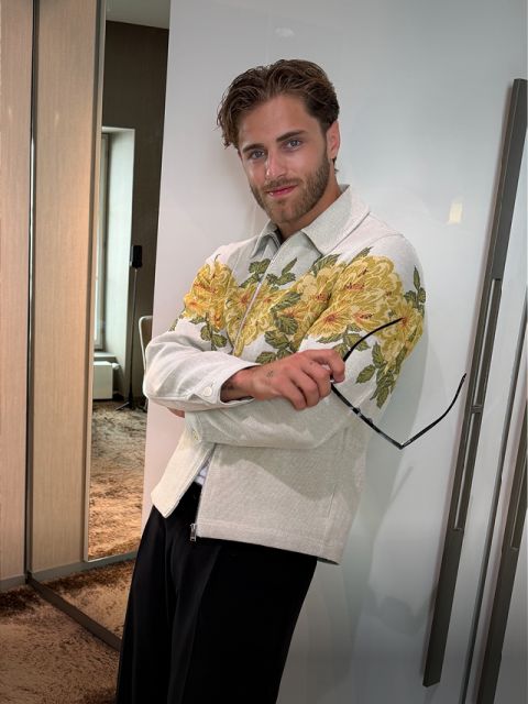 Ein bärtiger Mann trägt eine helle Jacke mit gelben Blumenmustern, hält eine Sonnenbrille in der Hand und steht in einem modernen Raum mit einem zotteligen Teppich.