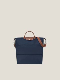 Eine rote Lederhandtasche mit zylindrischer Form steht auf einem weißen Hocker vor einem schlichten, neutralen Hintergrund. Die Tasche hat abgerundete Griffe und metallische Akzente.