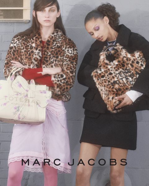 Eine Frau posiert im Freien mit einer braunen Marc-Jacobs-Tasche in der Hand, lehnt sich mit geneigtem Kopf zurück und trägt eine kastanienbraune Jacke und blaue Jeans. Auf dem Bild erscheinen die Worte "THE TOTE BAG MARC JACOBS" und "MARC JACOBS".