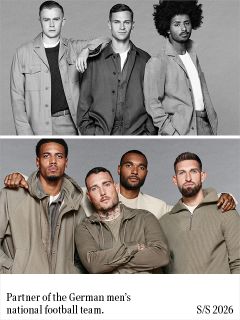 Ein Mann mit lockigem dunklem Haar, der einen schwarzen Blazer über einem grünen Rippstrickpullover trägt, steht mit verschränkten Armen vor einem einfarbigen grauen Hintergrund und blickt mit neutraler Miene geradeaus.