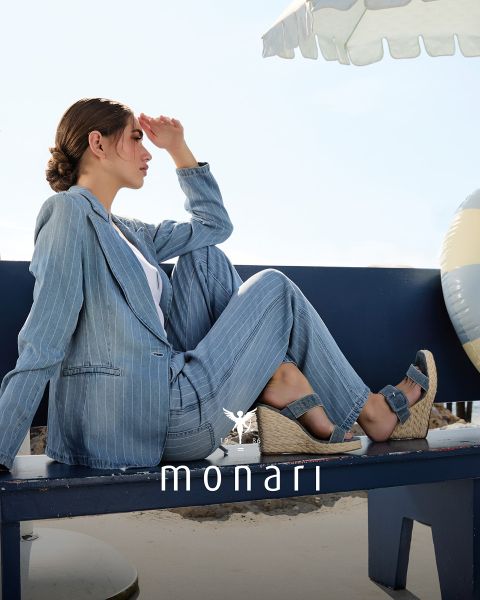 Eine lächelnde Frau, die einen beigen Mantel, ein gestreiftes Hemd, blaue Jeans und einen hellen Schal trägt, steht im Freien vor einem hohen Gebäude. Das Monari-Logo ist am unteren Rand des Bildes zu sehen.
