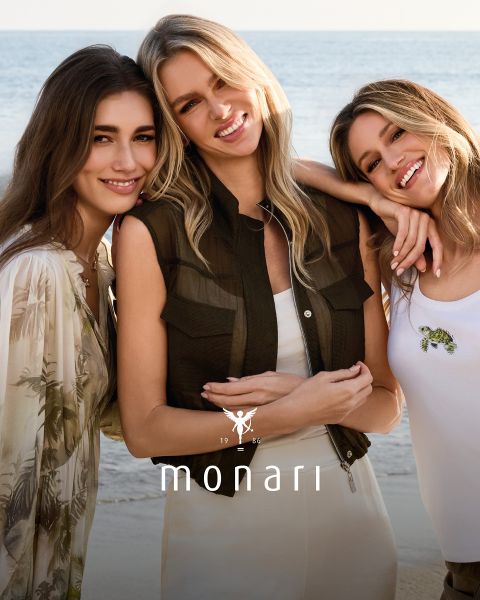 Eine lächelnde Frau, die einen beigen Mantel, ein gestreiftes Hemd, blaue Jeans und einen hellen Schal trägt, steht im Freien vor einem hohen Gebäude. Das Monari-Logo ist am unteren Rand des Bildes zu sehen.
