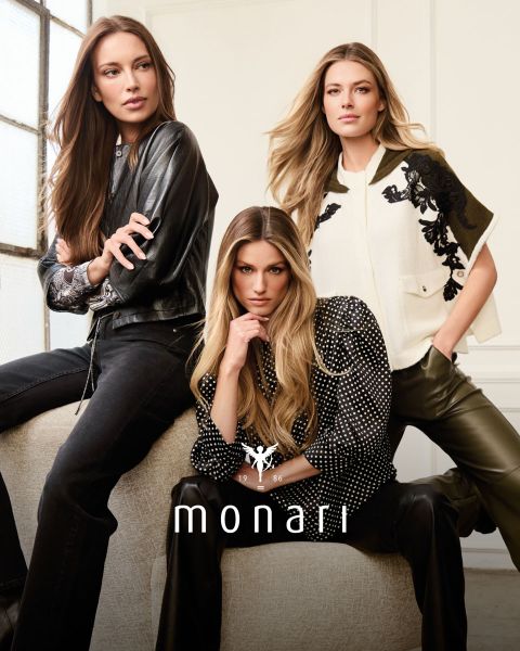 Eine lächelnde Frau, die einen beigen Mantel, ein gestreiftes Hemd, blaue Jeans und einen hellen Schal trägt, steht im Freien vor einem hohen Gebäude. Das Monari-Logo ist am unteren Rand des Bildes zu sehen.