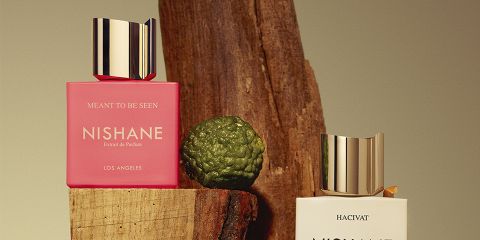 Eine Flasche Nishane-Parfüm mit goldenem Deckel ist von leuchtend roten, gelben und weißen Blumen umgeben. Daneben liegt eine Orangenschalenscheibe. Der Text lautet: „Enthüllen Sie Ihren eigenen Mythos mit NISHANE PARFUMES.“.