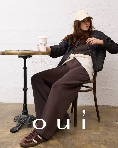 Eine Frau, die einen roten Pullover mit einem weißen Herz, blaue Jeans und eine weiße Mütze trägt, posiert vor einer hellen Wand. Am unteren Rand des Bildes steht in weißen Buchstaben das Wort "oui".