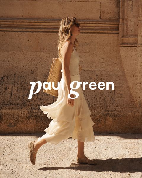 Eine Person mit langen roten Haaren, einer Sonnenbrille, einem hellgrünen Oberteil, beigen Hosen und blauen Turnschuhen sitzt auf felsigem Gelände unter blauem Himmel. Die Worte „Paul Green“ sind über das Bild gelegt.