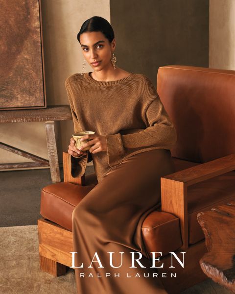 Eine Frau mit langem, gewelltem Haar lehnt auf einem weißen Sofa und trägt ein leuchtend rotes Oberteil und eine passende Hose. Warmes Sonnenlicht wirft Schatten auf die Szene. Am unteren Rand steht der Schriftzug "LAUREN RALPH LAUREN".