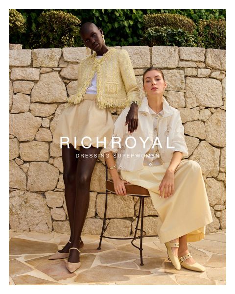 Eine Frau in hellblauem Hemd und grauer Hose hält sich eine Schneekugel vor ein Auge. Sie posiert in einem Raum mit Holzwänden und einem Skiplakat und trägt große Ohrringe. Der Text lautet "RICHROYAL DRESSING SUPERWOMEN".