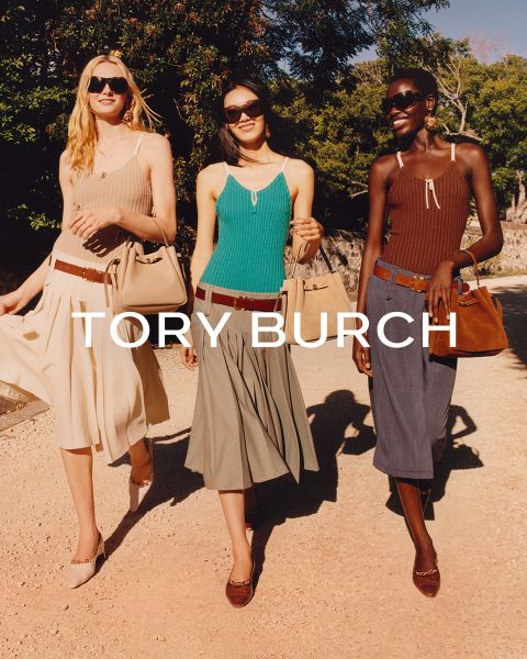 Eine Frau in einem schimmernden Oberteil und einem grünen Rock sitzt neben grünen Geschenkkartons und hält eine geblümte Tory-Burch-Tasche in der Hand; hinter ihr steht ein geschmückter Weihnachtsbaum.